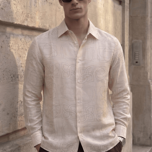A man wearing a light beige long-sleeve button-up shirt with a subtle tonal geometric pattern, dark sunglasses, and dark trousers, standing against a textured stone wall.