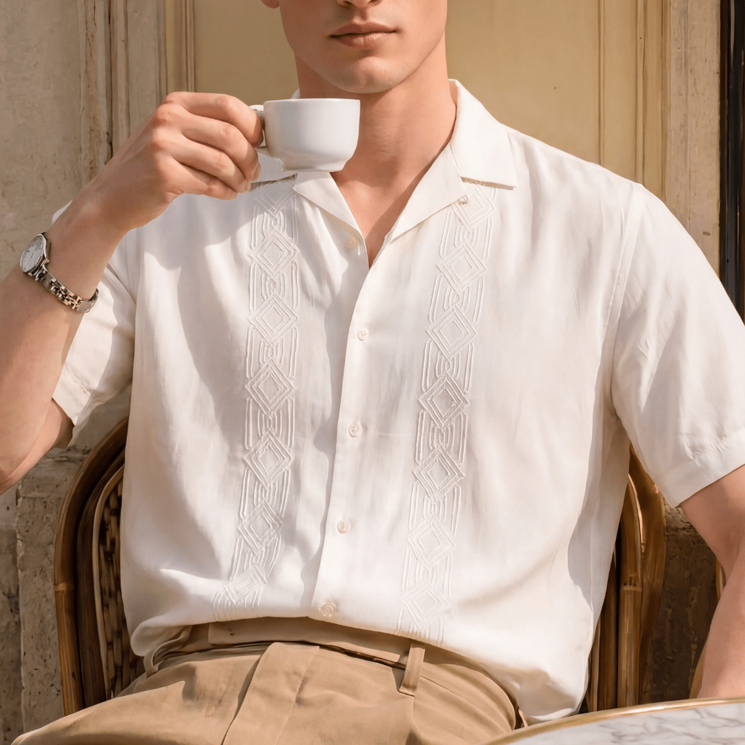 A man wearing a short-sleeve white button-up shirt with vertical embroidered patterns and khaki pants, holding a white coffee cup in front of his face. He is wearing dark sunglasses and a silver watch, seated in a wicker chair against a beige stone wall.
