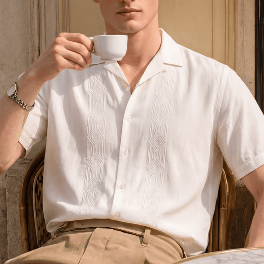 A man wearing a short-sleeve white button-up shirt with vertical embroidered patterns and khaki pants, holding a white coffee cup in front of his face. He is wearing dark sunglasses and a silver watch, seated in a wicker chair against a beige stone wall.