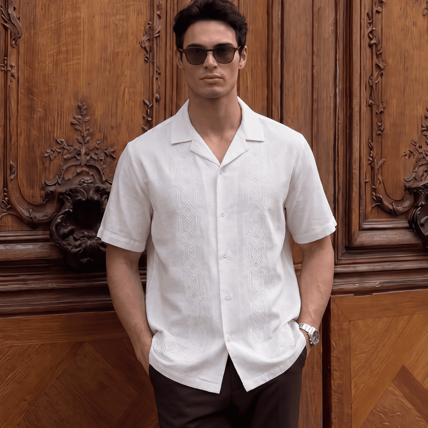 A man wearing a white short-sleeve button-up shirt and dark brown trousers stands in front of an ornate wooden door, wearing dark sunglasses and a wristwatch.