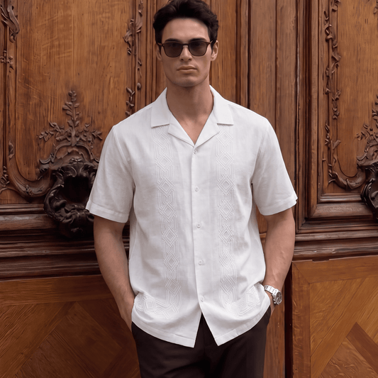 A man wearing a white short-sleeve button-up shirt and dark brown trousers stands in front of an ornate wooden door, wearing dark sunglasses and a wristwatch.