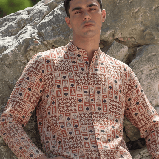 A man wearing a long-sleeve button-up shirt with a geometric and tribal pattern in terracotta red, beige, and black, posed outdoors against a large rock.