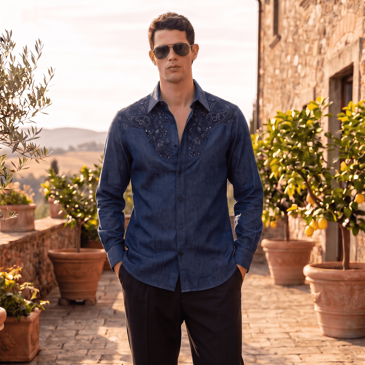 A man wearing a dark blue denim western-style button-down shirt with black embroidered yoke detailing, black pants, and sunglasses, standing on a stone patio with potted citrus trees and a rustic stone building in the background.