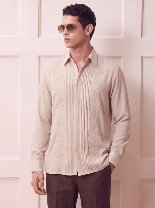 A man wearing a light beige long-sleeve button-up shirt with vertical pleats and embroidered details, paired with dark brown trousers and gold-framed sunglasses, standing against a light pink paneled wall.