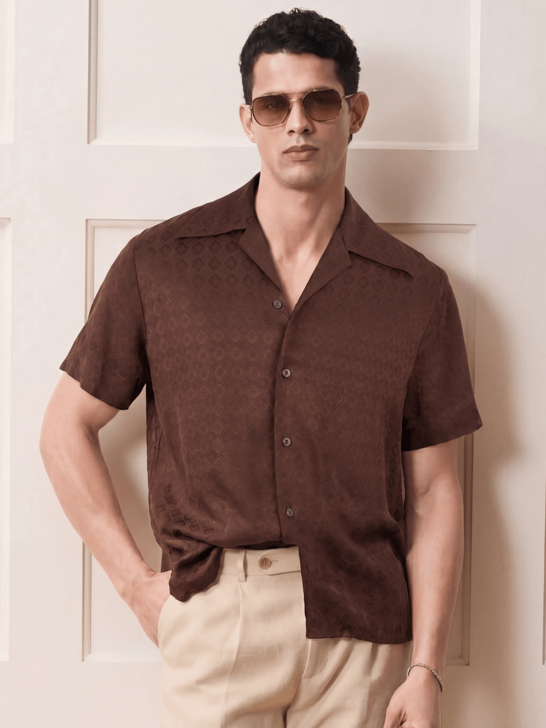 A man wearing a brown short-sleeve button-up shirt with a subtle tonal geometric pattern, paired with beige trousers and gold-tinted sunglasses, standing against a cream-colored paneled wall.