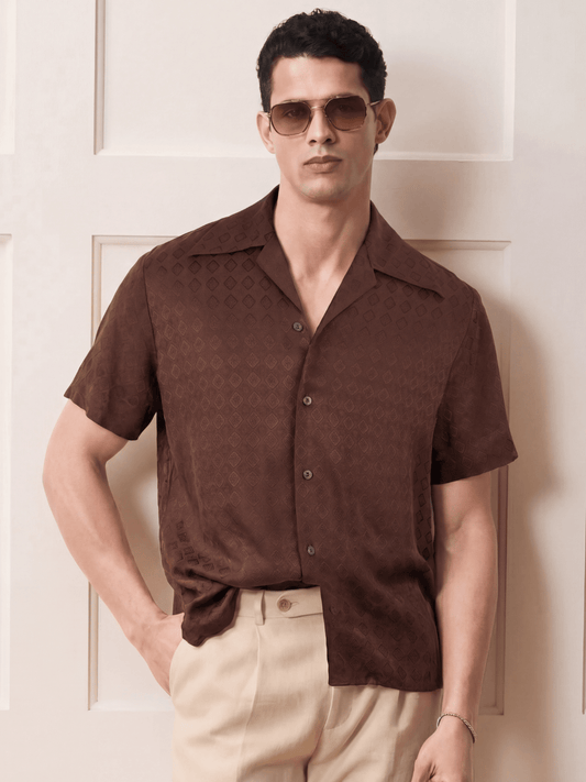 A man wearing a brown short-sleeve button-up shirt with a subtle tonal geometric pattern, paired with beige trousers and gold-tinted sunglasses, standing against a cream-colored paneled wall.