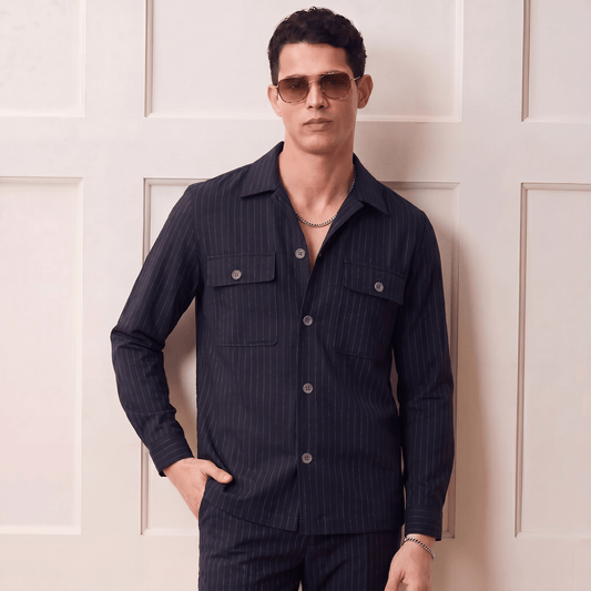 Man wearing a dark navy pinstripe shirt and pants with sunglasses against a light-colored wall.