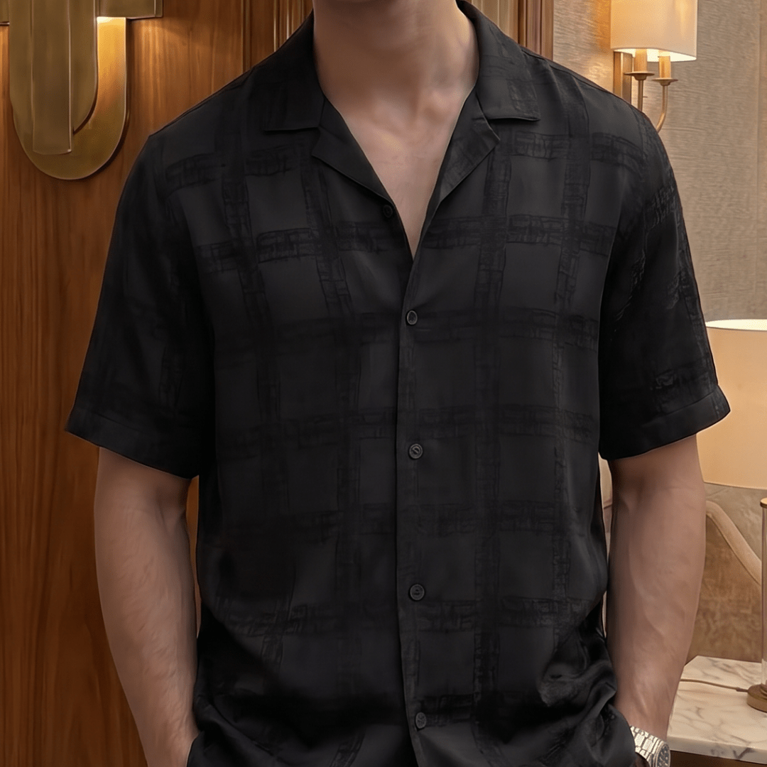A man wearing a black short-sleeve button-up shirt with a subtle plaid pattern, styled with dark sunglasses in an indoor setting with warm lighting.