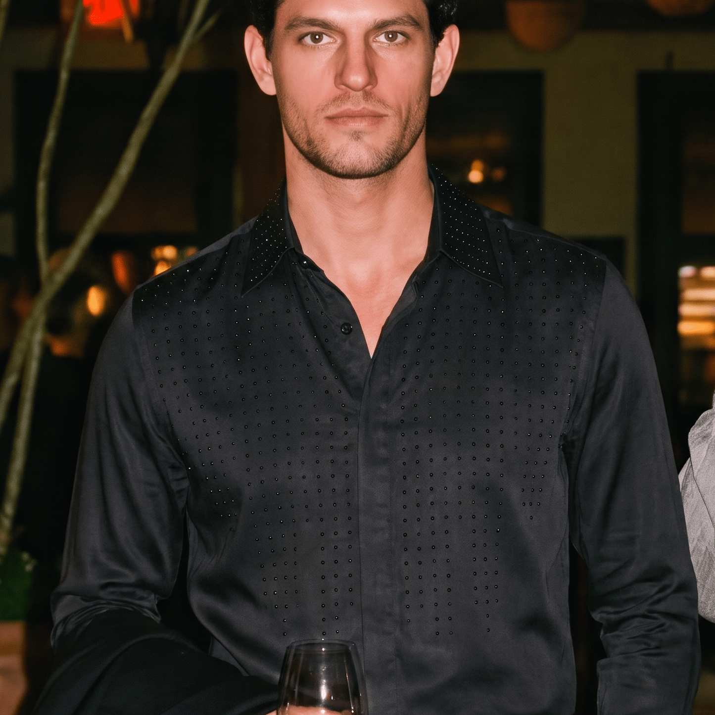 A man wearing a black long-sleeve button-down shirt with a subtle textured pattern, holding a glass of red wine in a dimly lit indoor setting.