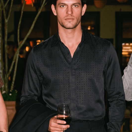 A man wearing a black long-sleeve button-down shirt with a subtle textured pattern, holding a glass of red wine in a dimly lit indoor setting.