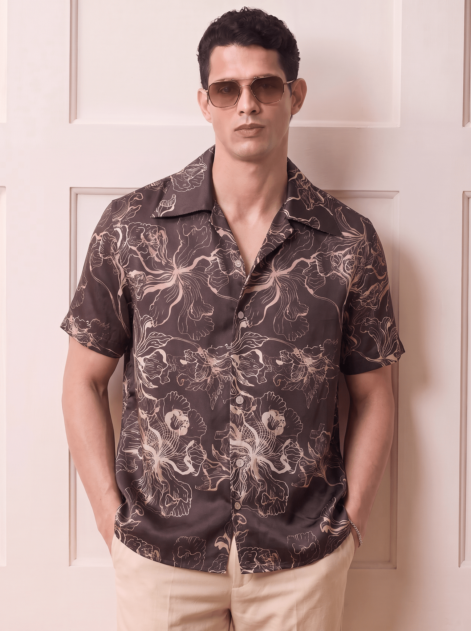 A man wearing a dark brown short-sleeve button-up shirt with a beige and white floral pattern, paired with beige pants and gold-tinted sunglasses, standing in front of a cream-colored paneled door.