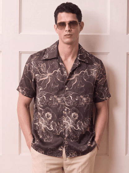 A man wearing a dark brown short-sleeve button-up shirt with a beige and white floral pattern, paired with beige pants and gold-tinted sunglasses, standing in front of a cream-colored paneled door.