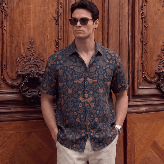 A man wearing a short-sleeve button-up shirt with a dark blue and brown floral pattern, paired with light beige trousers, standing in front of an ornate wooden door.