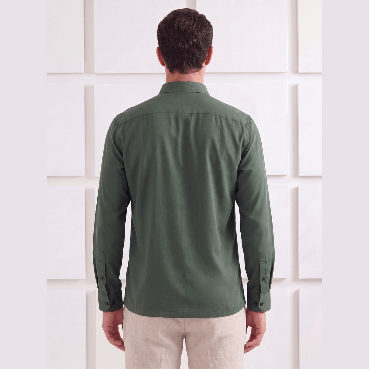 A man viewed from behind wearing a dark olive green long-sleeve button-up shirt and light beige trousers, standing against a white textured wall.