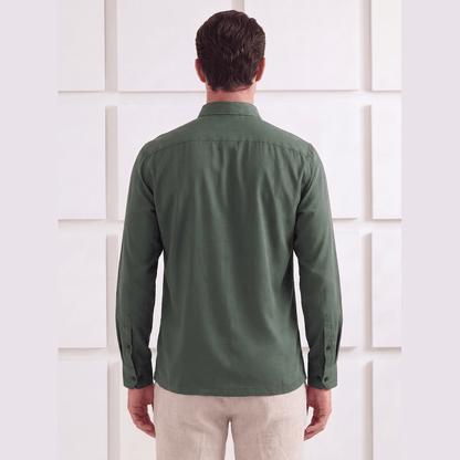 A man viewed from behind wearing a dark olive green long-sleeve button-up shirt and light beige trousers, standing against a white textured wall.