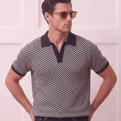 A man wearing a short-sleeve polo shirt with a black and white chevron pattern, dark navy collar and sleeve cuffs, and gold-framed sunglasses, standing against a light pink paneled wall.