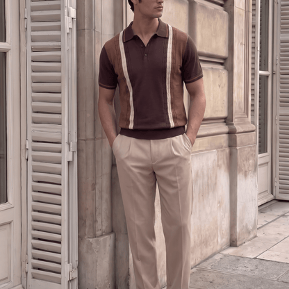 A man wearing a brown and beige striped knit polo shirt, beige trousers, and white sneakers stands against a stone building wall with shuttered windows.