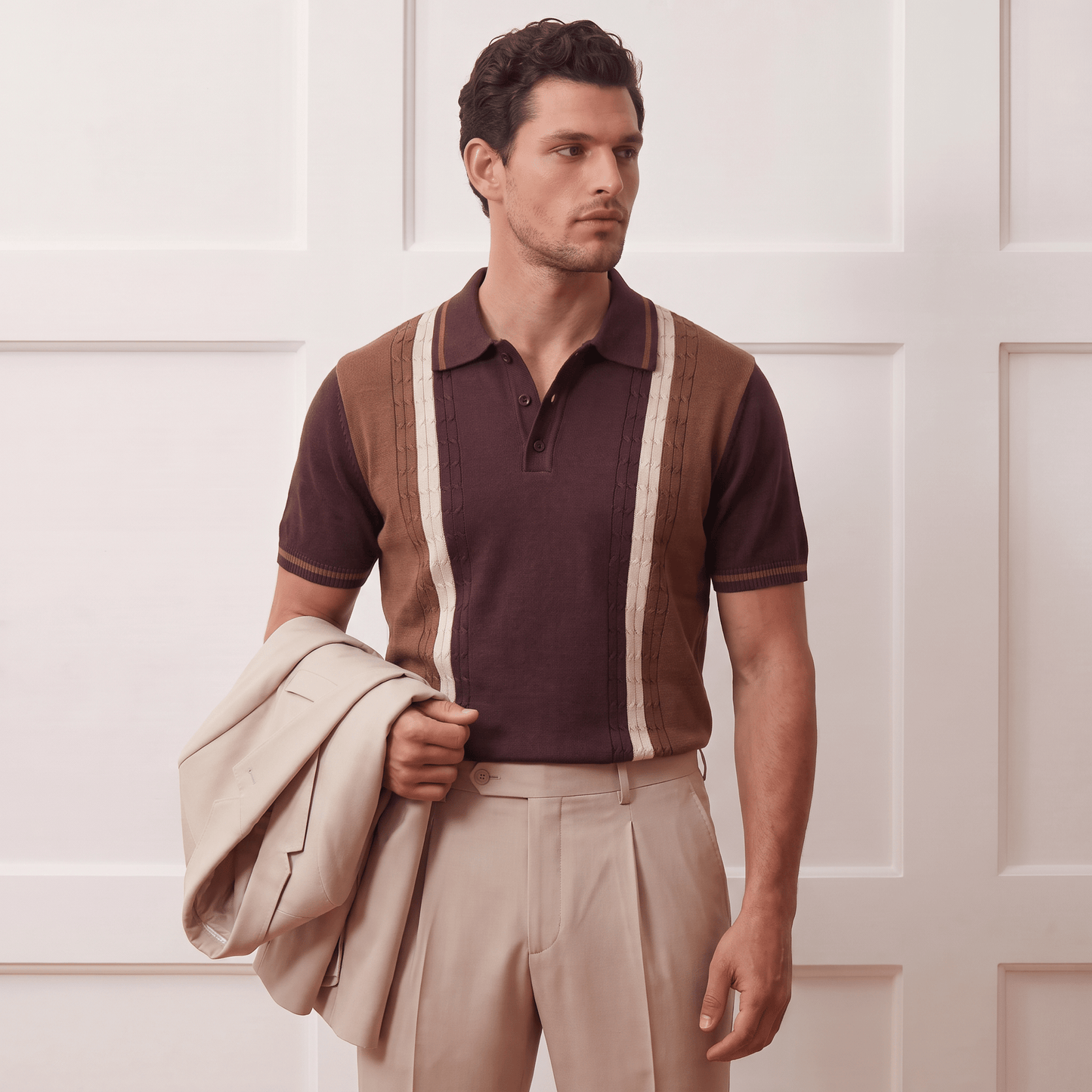 A man wearing a brown and beige knit polo sweater vest over a dark brown polo shirt, paired with beige trousers and holding a light beige blazer, standing against a white paneled wall.