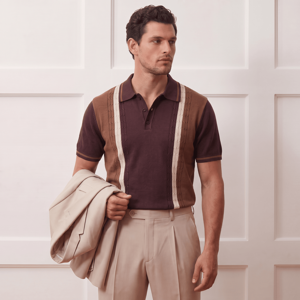 A man wearing a brown and beige knit polo sweater vest over a dark brown polo shirt, paired with beige trousers and holding a light beige blazer, standing against a white paneled wall.