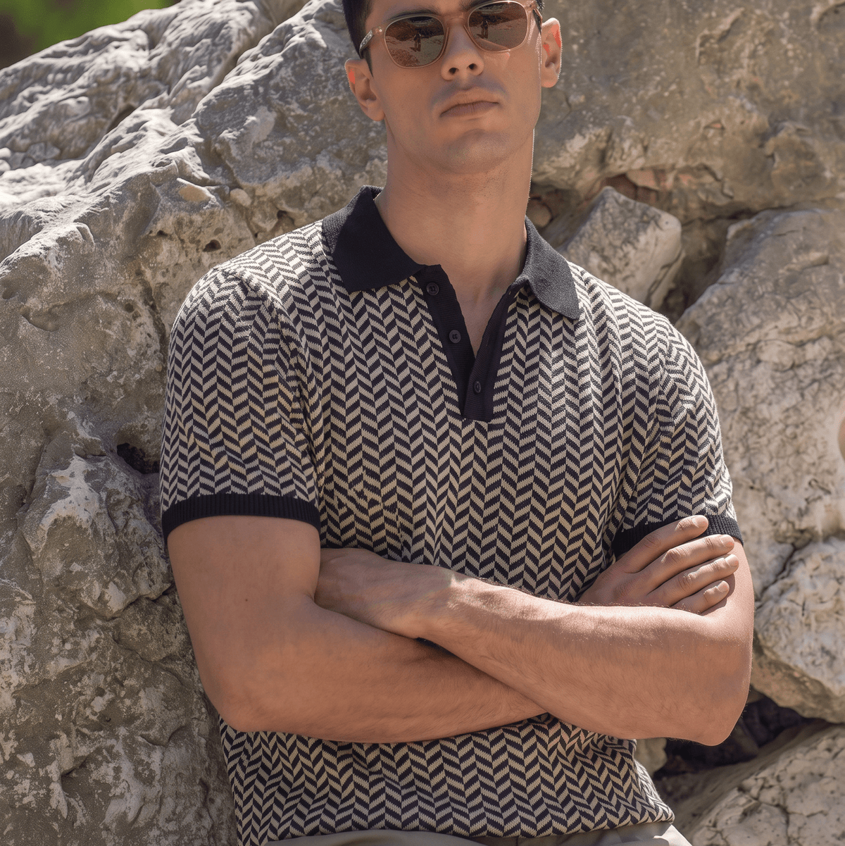 A man wearing a short-sleeve polo shirt with a brown and beige chevron pattern and black collar/cuffs, sitting outdoors against a large rock.