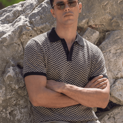 A man wearing a short-sleeve polo shirt with a brown and beige chevron pattern and black collar/cuffs, sitting outdoors against a large rock.