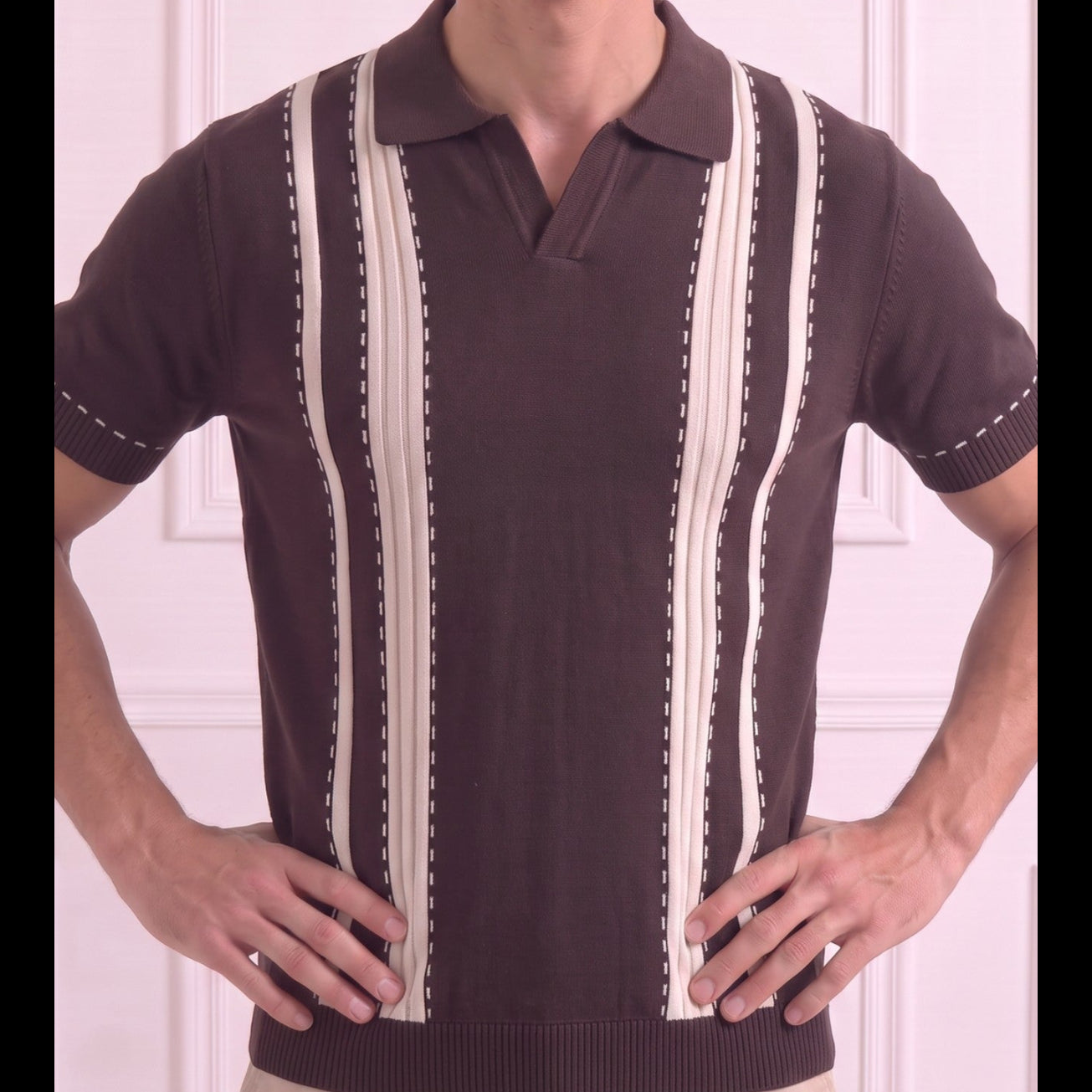 A man wearing a dark brown short-sleeve polo sweater with vertical cream-colored stripes and dotted stitching details, paired with beige pants, standing against a light pink paneled wall.