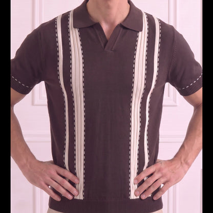 A man wearing a dark brown short-sleeve polo sweater with vertical cream-colored stripes and dotted stitching details, paired with beige pants, standing against a light pink paneled wall.
