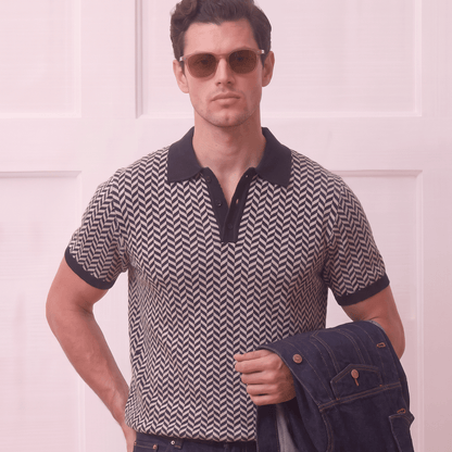 A man wearing a short-sleeve polo shirt with a black and beige chevron pattern, dark navy jeans, and sunglasses, holding a dark denim jacket against a light pink paneled background.