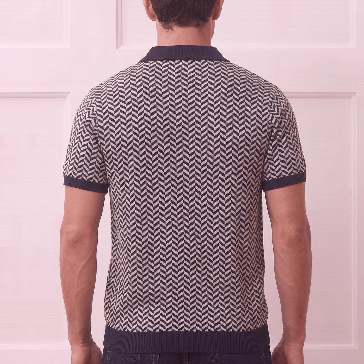 A man viewed from behind wearing a short-sleeve knit polo shirt with a navy and beige chevron pattern, paired with dark blue jeans, set against a light pink paneled wall.