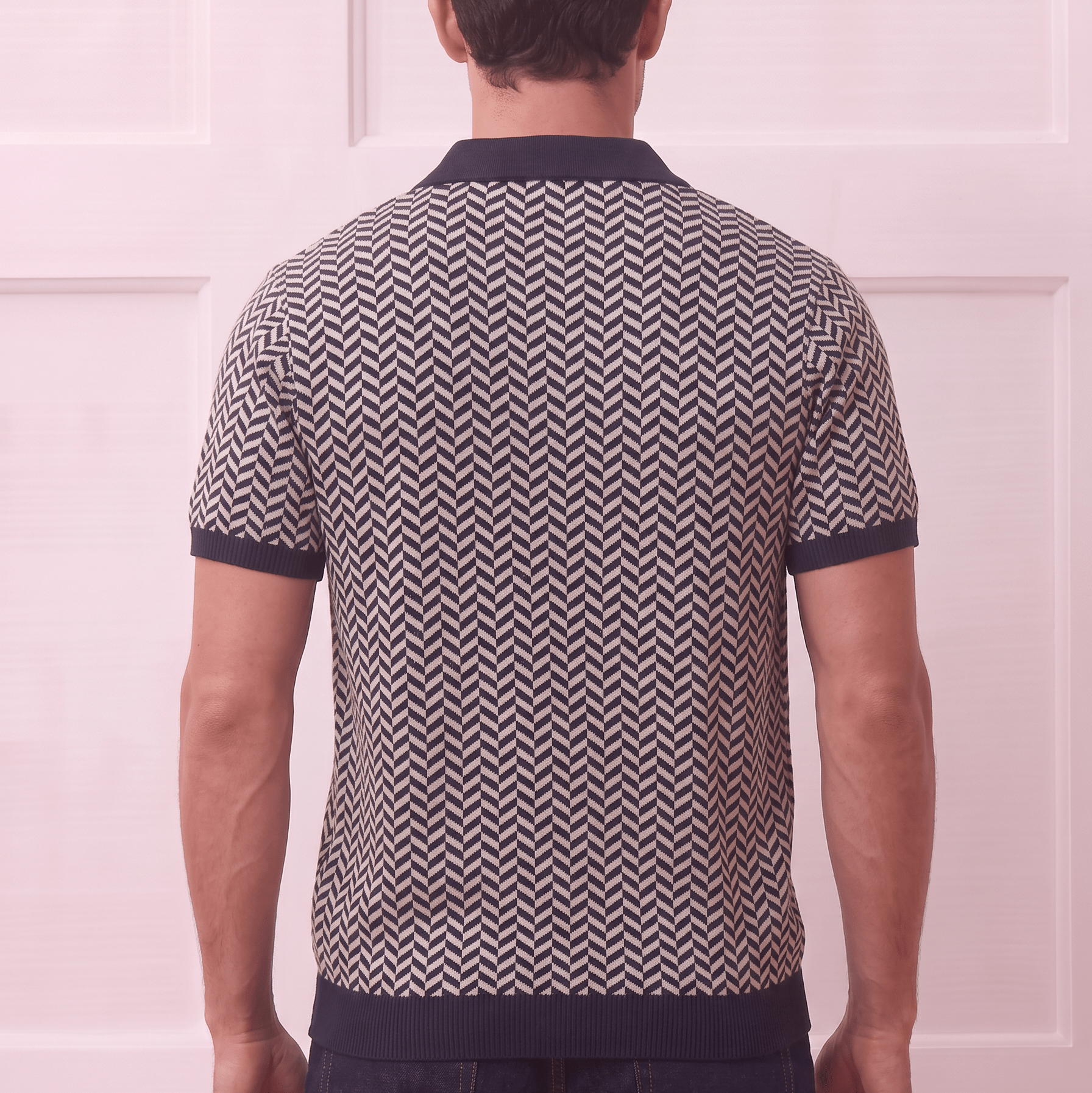 A man viewed from behind wearing a short-sleeve knit polo shirt with a navy and beige chevron pattern, paired with dark blue jeans, set against a light pink paneled wall.