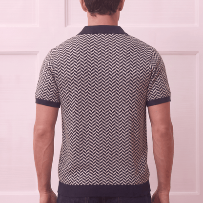 A man viewed from behind wearing a short-sleeve knit polo shirt with a navy and beige chevron pattern, paired with dark blue jeans, set against a light pink paneled wall.