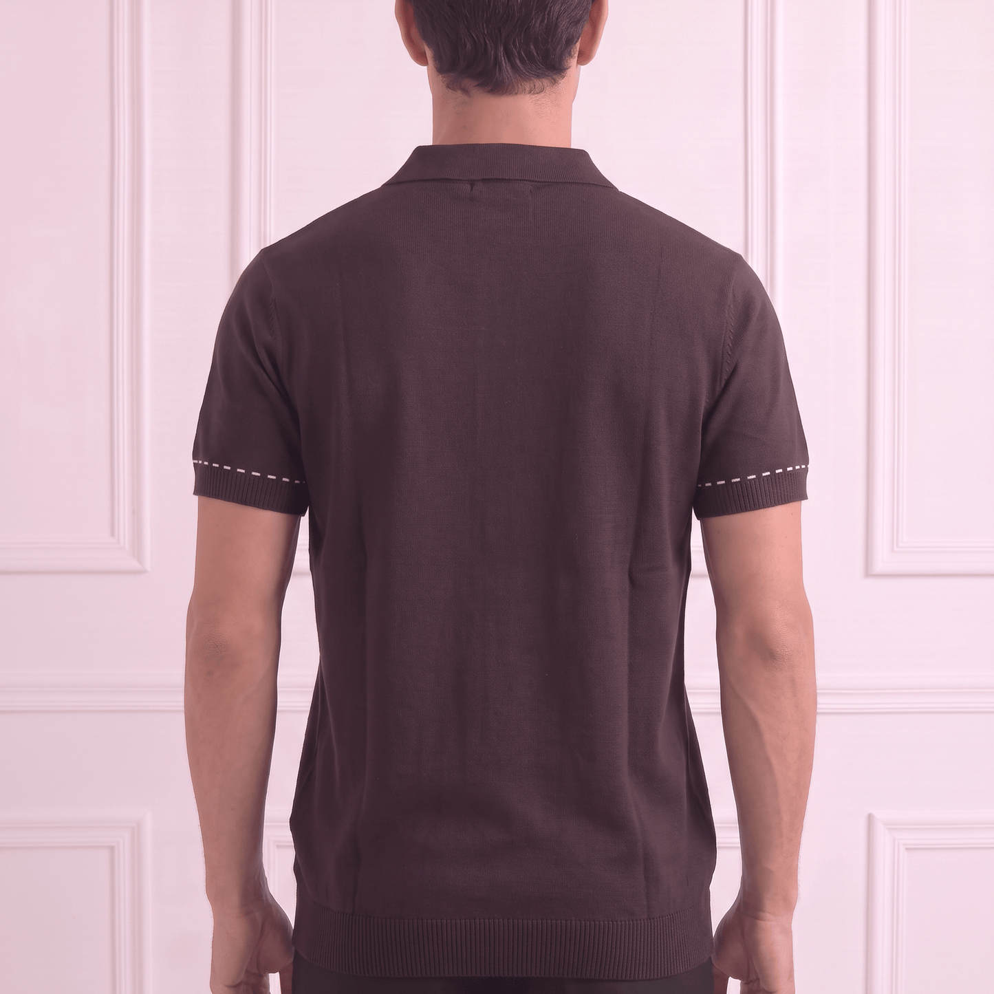 A man viewed from behind wearing a dark brown short-sleeve knit polo shirt with white contrast stitching on the sleeve cuffs, paired with black trousers, standing against a light pink paneled wall.