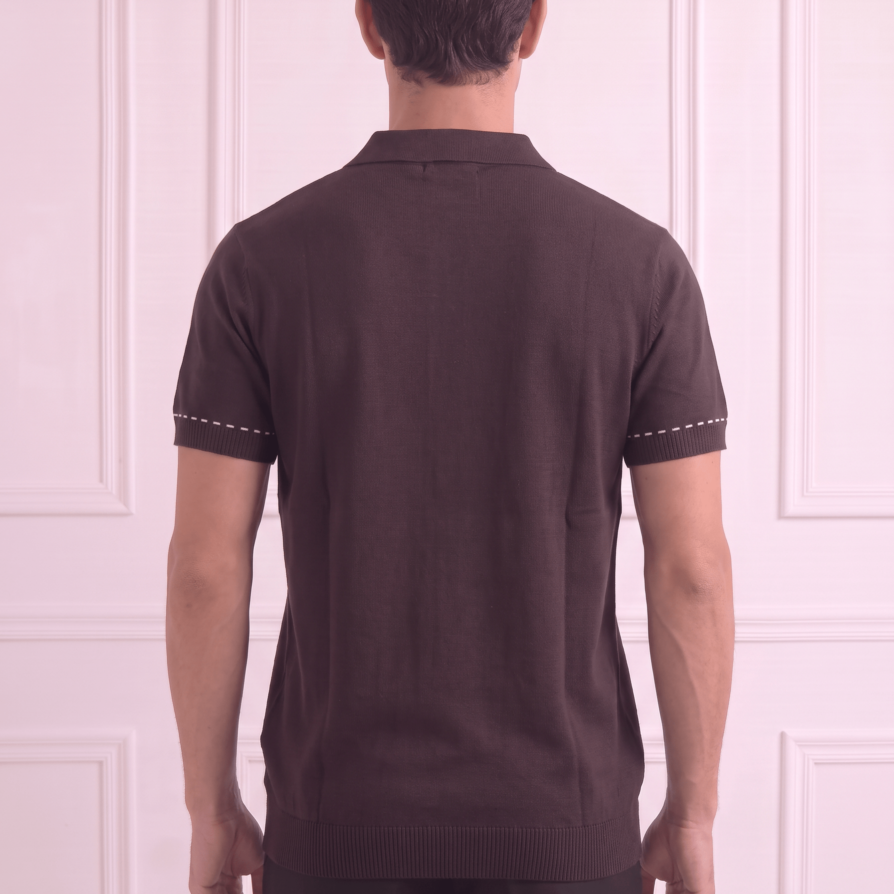 A man viewed from behind wearing a dark brown short-sleeve knit polo shirt with white contrast stitching on the sleeve cuffs, paired with black trousers, standing against a light pink paneled wall.
