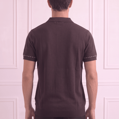 A man viewed from behind wearing a dark brown short-sleeve knit polo shirt with white contrast stitching on the sleeve cuffs, paired with black trousers, standing against a light pink paneled wall.