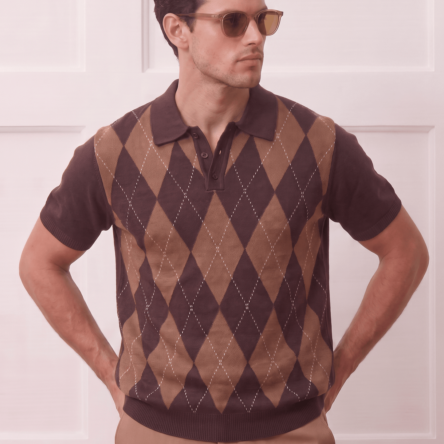 A man wearing a brown and tan argyle knit polo sweater vest over a dark brown short-sleeve polo shirt, paired with tan trousers and gold-framed sunglasses, standing against a white paneled wall.