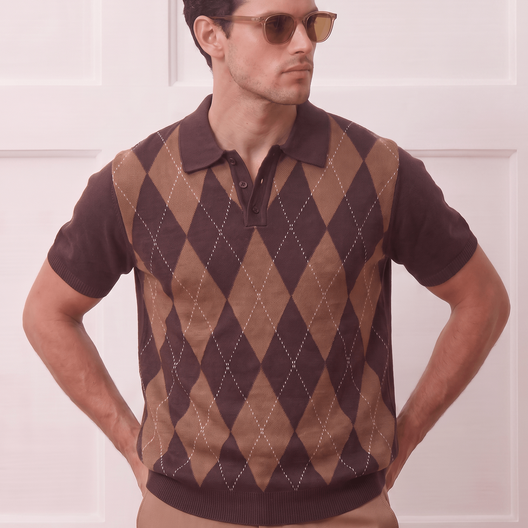 A man wearing a brown and tan argyle knit polo sweater vest over a dark brown short-sleeve polo shirt, paired with tan trousers and gold-framed sunglasses, standing against a white paneled wall.