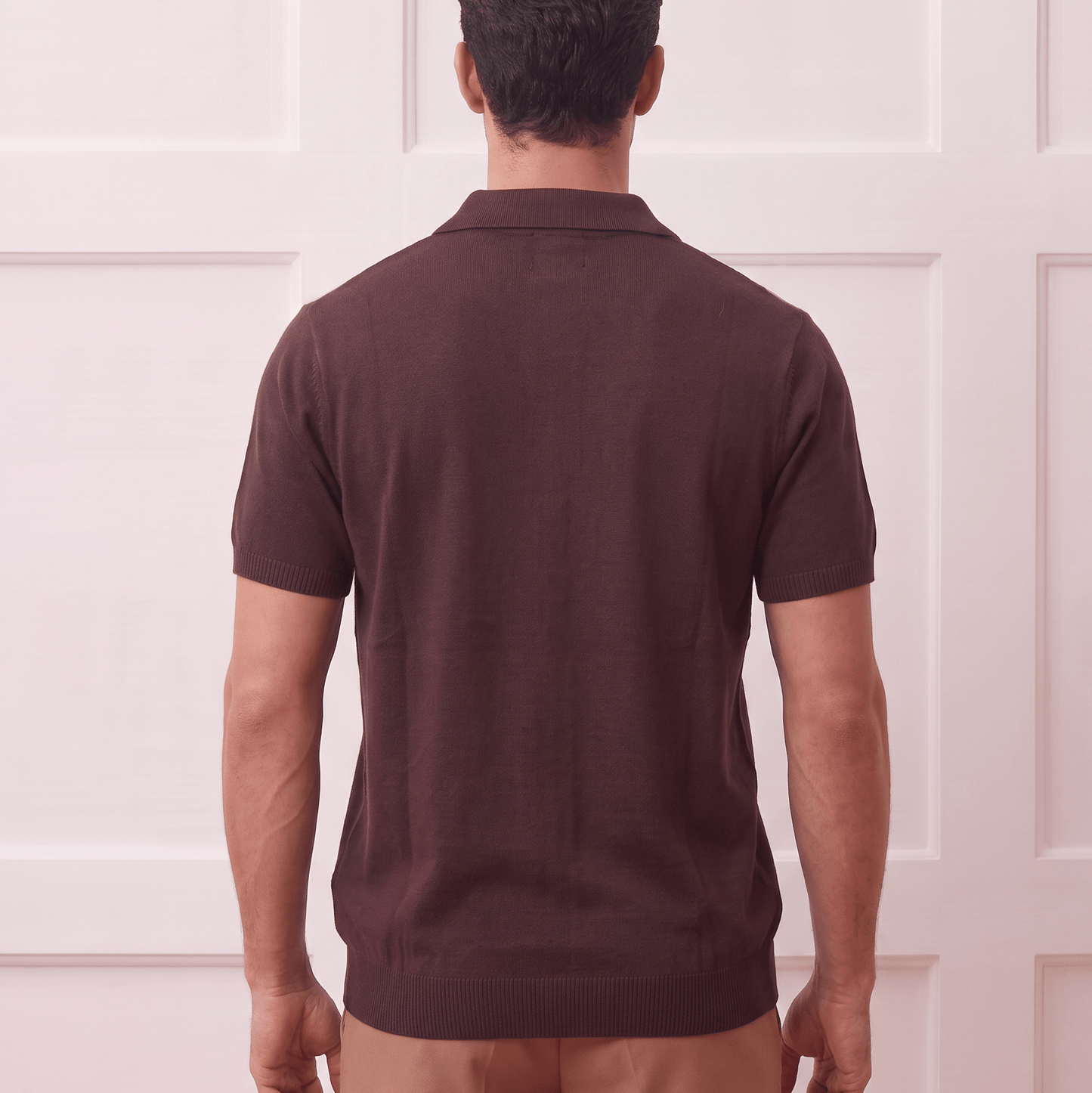 A man viewed from behind wearing a dark brown short-sleeve polo shirt and tan trousers, standing against a white paneled wall.