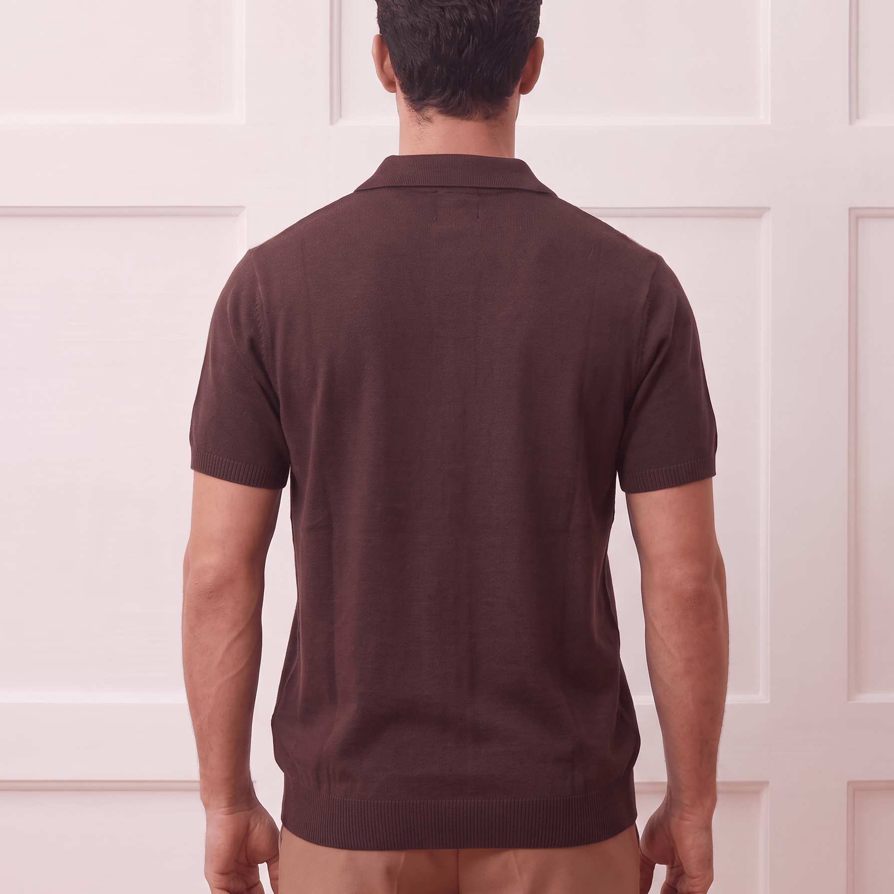 A man viewed from behind wearing a dark brown short-sleeve polo shirt and tan trousers, standing against a white paneled wall.