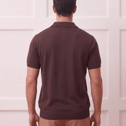 A man viewed from behind wearing a dark brown short-sleeve polo shirt and tan trousers, standing against a white paneled wall.