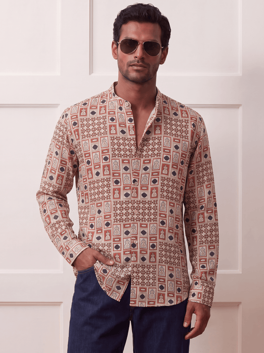 A man wearing a long-sleeve button-up shirt with a beige, brown, and red geometric pattern, paired with dark blue trousers and gold aviator sunglasses, standing against a white paneled wall.