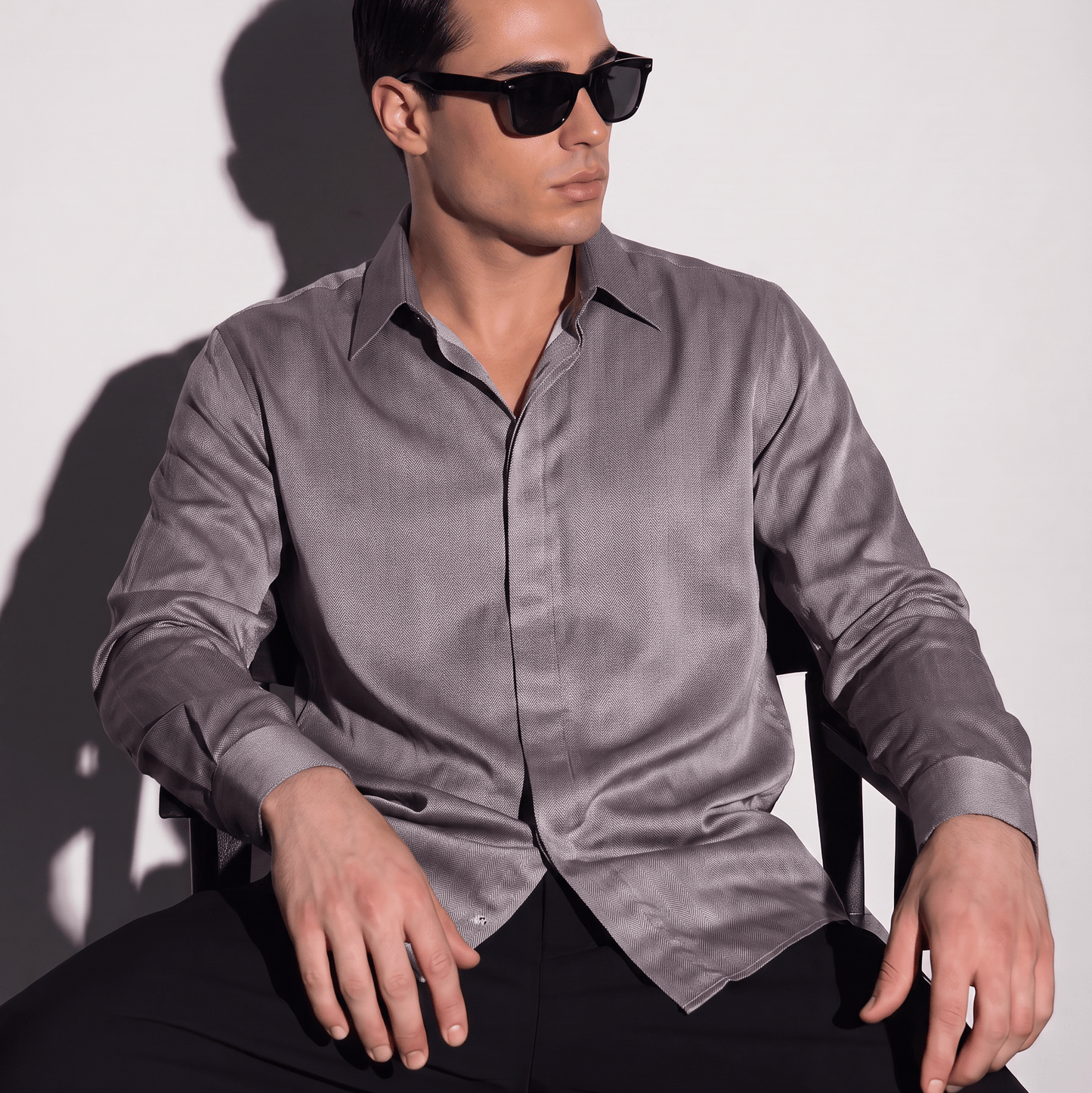 A man wearing a long-sleeve satin-like button-down shirt in a muted grayish purple and black sunglasses, seated against a plain light gray wall with a hard shadow.