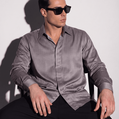 A man wearing a long-sleeve satin-like button-down shirt in a muted grayish purple and black sunglasses, seated against a plain light gray wall with a hard shadow.