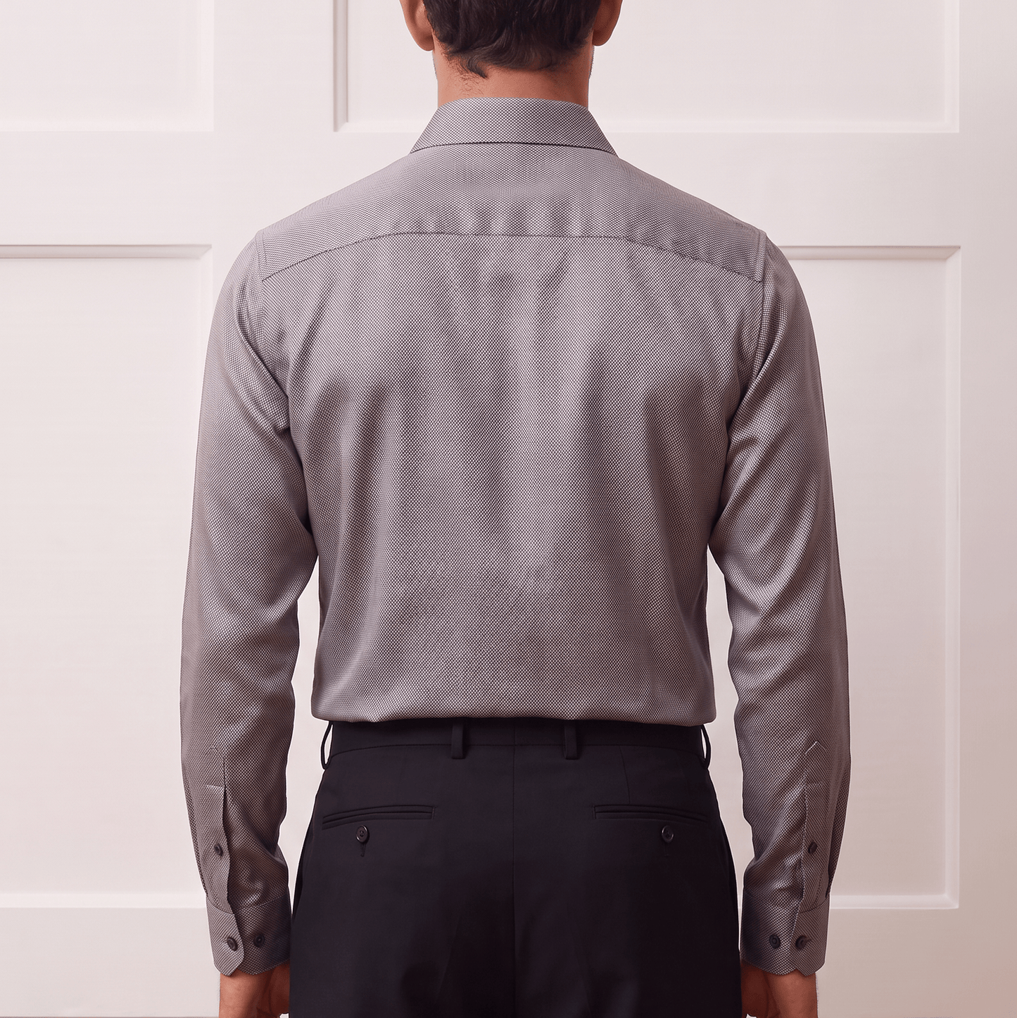 A man viewed from behind wearing a light gray long-sleeve button-down shirt and dark trousers, standing against a white paneled wall.