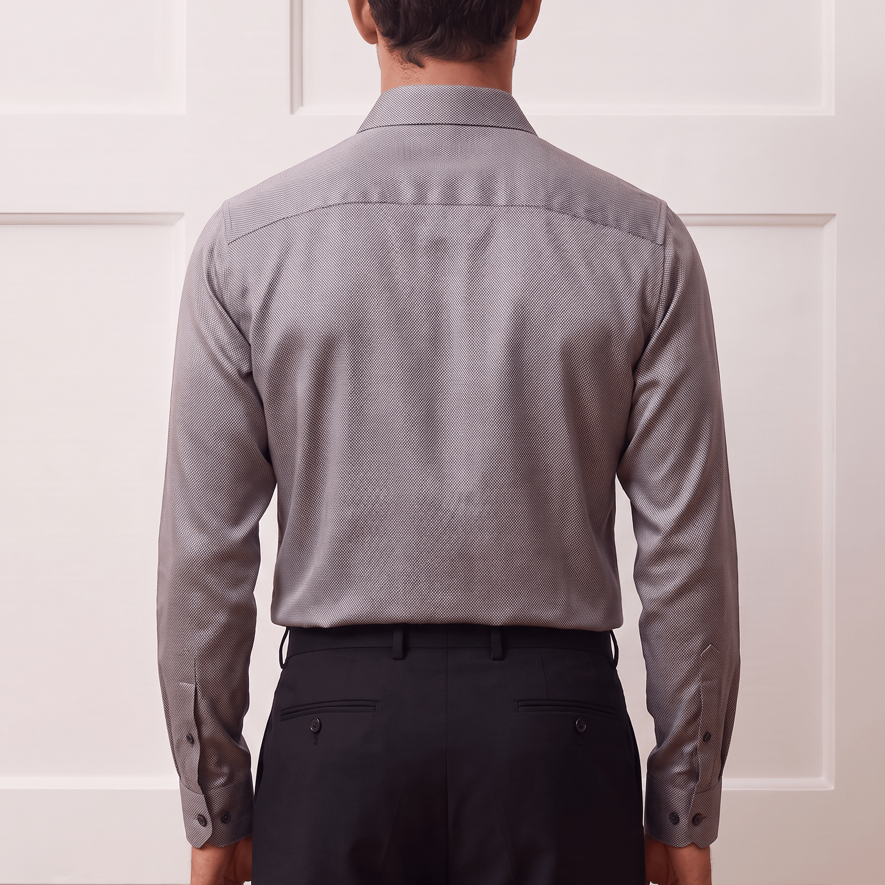 A man viewed from behind wearing a light gray long-sleeve button-down shirt and dark trousers, standing against a white paneled wall.