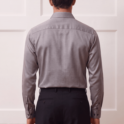 A man viewed from behind wearing a light gray long-sleeve button-down shirt and dark trousers, standing against a white paneled wall.