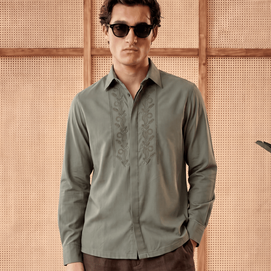 A man wearing a muted sage green long-sleeve button-up shirt with tonal embroidery on the chest, paired with dark brown trousers and black sunglasses, standing against a beige woven wall.