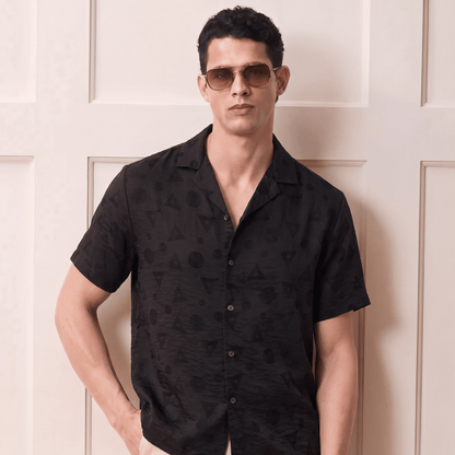 A man wearing a black short-sleeve button-up shirt with a subtle tonal pattern, paired with beige pants and sunglasses, standing against a cream-colored paneled wall.